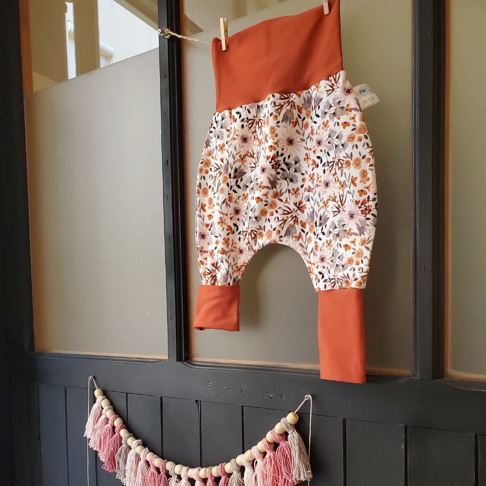 Pantalon évolutif blanc avec des fleurs beiges et oranges et des bandes oranges en haut et en bas accroché sur un fil dans une boutique