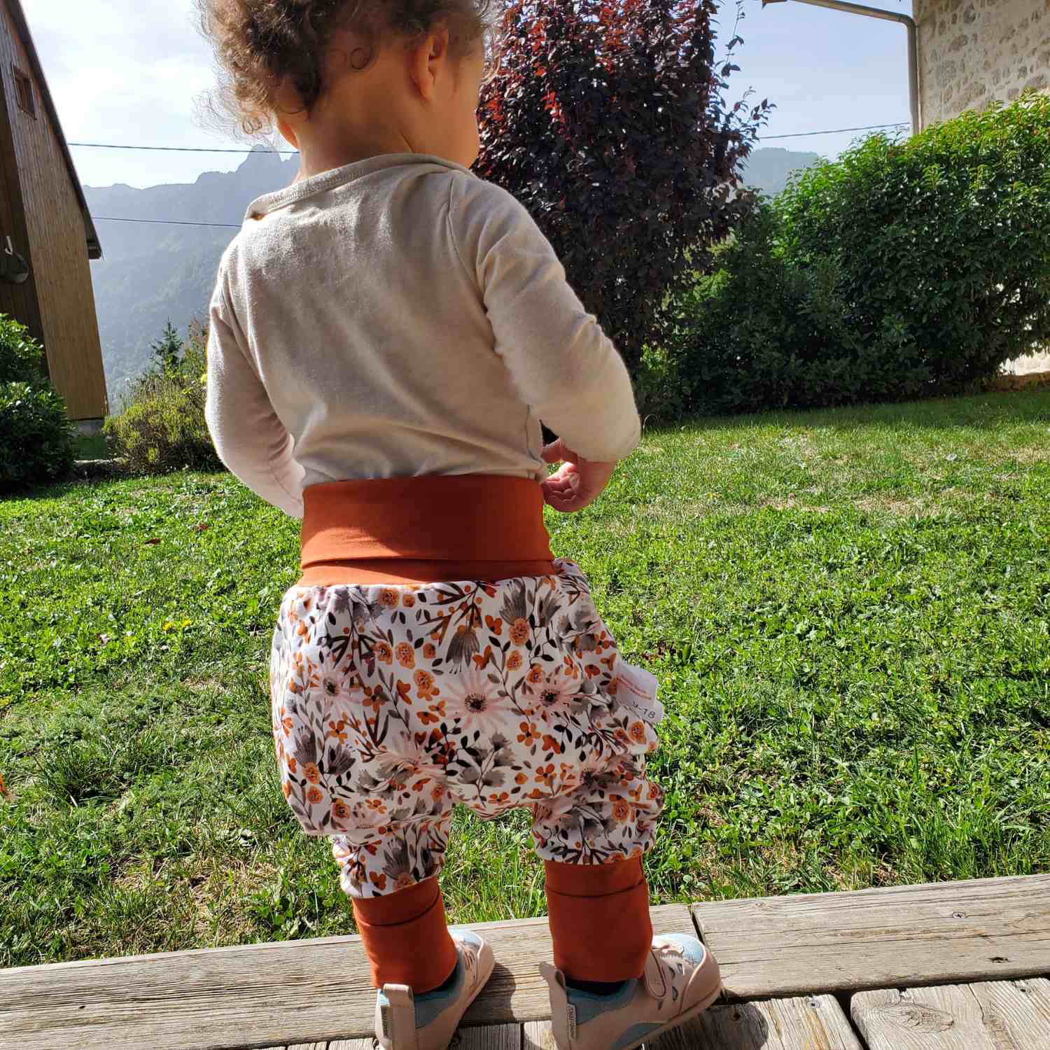 Bébé de dos dans un jardin portant un pantalon évolutif blanc avec des fleurs beiges et oranges